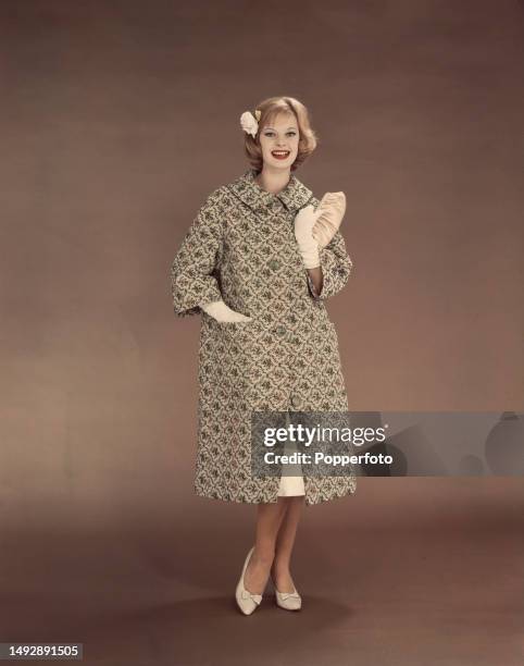 Posed studio portrait of a female fashion model wearing a tapestry swing style coat over a white dress, she wears white gloves along with cream...