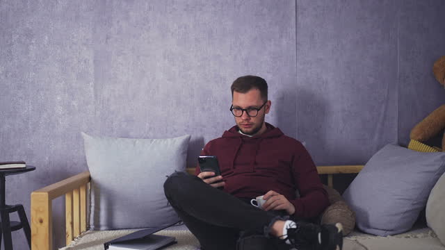 Young Bearded Male Spending Free Time with Coffee and Phone in Cozy Living Room