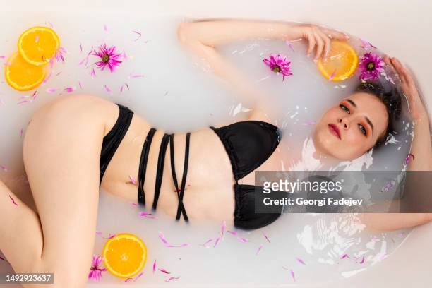 tender girl lying in a bath half in water filled with fruit orange slices and flower petals. beauty and skin care concept. top view. - woman bath tub wet hair stock pictures, royalty-free photos & images