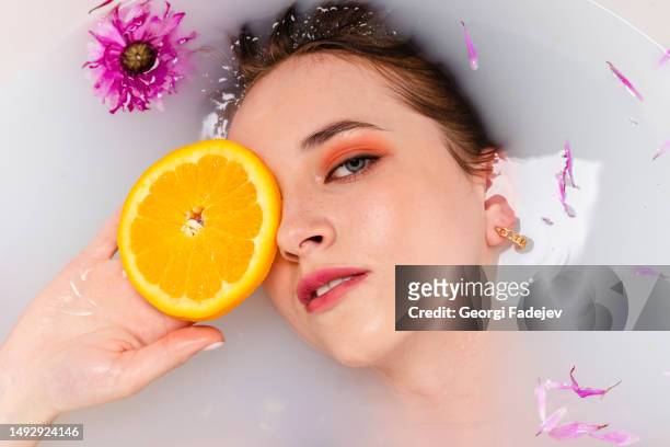 tender girl lying in a bath half in water filled with fruit orange slices and flower petals. beauty and skin care concept. top view. head shot. - badewanne mit obst stock-fotos und bilder
