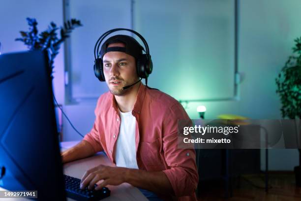 jeune bel homme avec chapeau et casque jouant à des jeux vidéo sur pc de bureau à la maison - joueur de jeux informatiques photos et images de collection