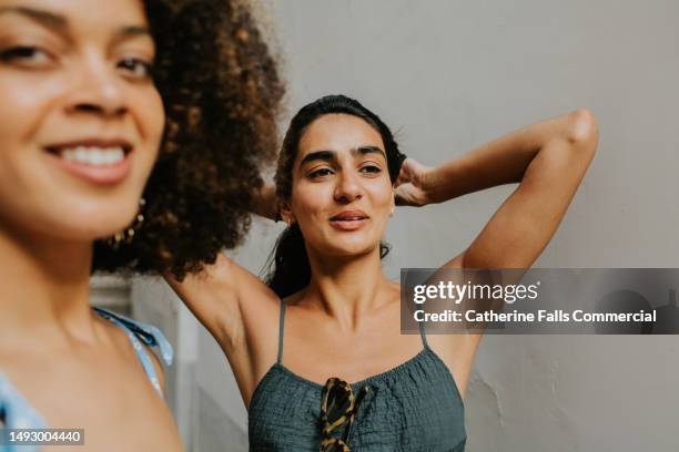 candid image of two woman in front of a plain background. one woman reaches her arms over her head. - armpit hair woman stock pictures, royalty-free photos & images