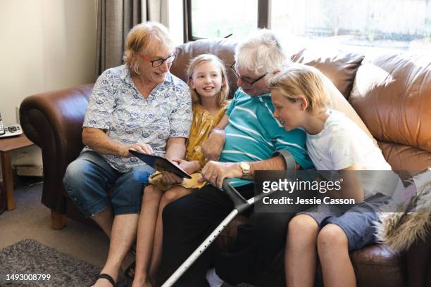 senior man holding digital tablet sitting on sofa with senior wife and grandchildren - ageing population stock pictures, royalty-free photos & images