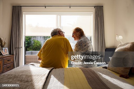 Back shot of senior couple sitting on edge of bed, in front of window Back shot of senior couple sitting on edge of bed, in front of window