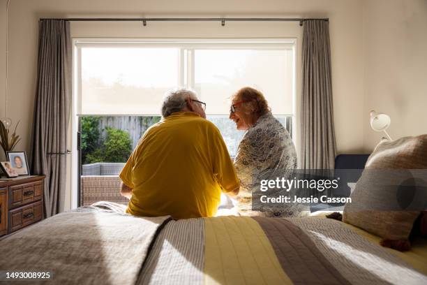 back shot of senior couple sitting on edge of bed, in front of window - demens bildbanksfoton och bilder