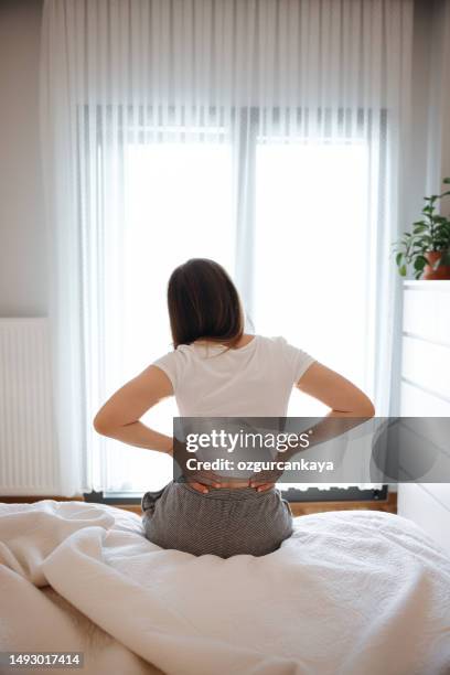 young woman sitting on bed and suffering from back pain stock - human joint stock pictures, royalty-free photos & images