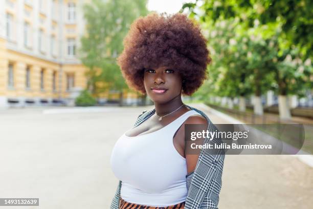 beautiful young black woman standing and looking at camera with building and blooming chestnut trees in background - charming stockfoto's en -beelden