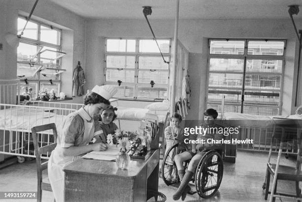 Nurse writes at her desk in a hospital children's ward while two girls and a boy in a wheelchair look on, October 1954. Original Publication: Picture...