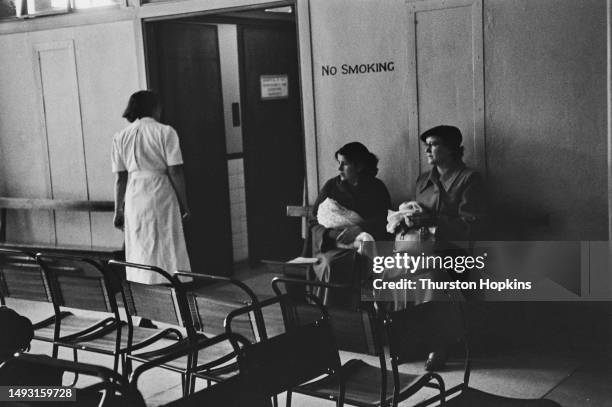 Two women sit in a hospital waiting room, one carrying a baby, while a nurse stands nearby, October 1954. Original Publication: Picture Post - 7333 -...