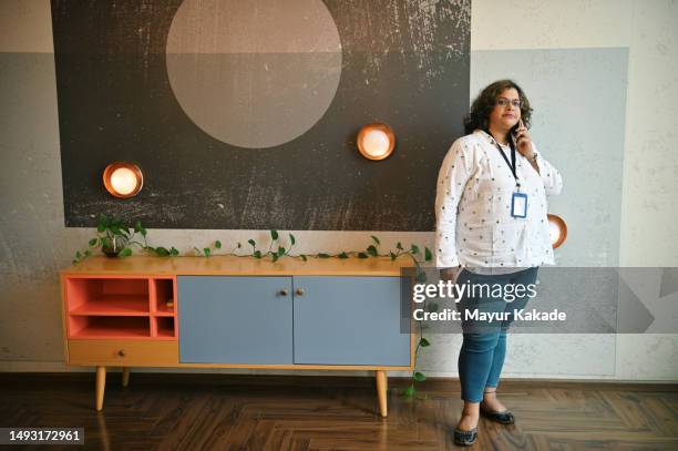 a mid-adult woman confidently speaking on the mobile phone standing in a co-working office - co op stock pictures, royalty-free photos & images