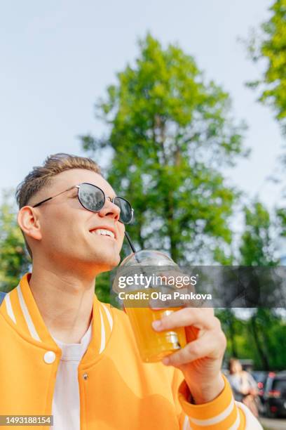 cheerful young man sporting a yellow jacket and sunglasses, stroll through the city while indulging in fresh orange juice. - orange coat foto e immagini stock