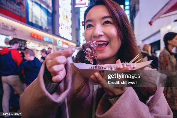 a female tourist in osaka is holding a freshly made japanese traditional street food called takoyaki (octopus balls) as she explores the city center streets. we can see her hand holding the piping hot takoyaki skewer, wafting with a tantalizing aroma. - takoyaki stock pictures, royalty-free photos & images