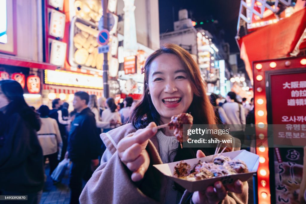 A female tourist in Osaka is holding a freshly made Japanese traditional street food called Takoyaki (octopus balls) as she explores the city center streets. We can see her hand holding the piping hot Takoyaki skewer, wafting with a tantalizing aroma.