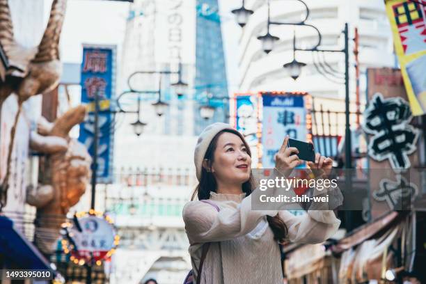 eine asiatische frau besichtigt tsutenkaku in osaka, japan. mit ihrem smartphone macht sie fotos von tsutenkaku und der umliegenden landschaft. - osaka shinsekai food stock-fotos und bilder