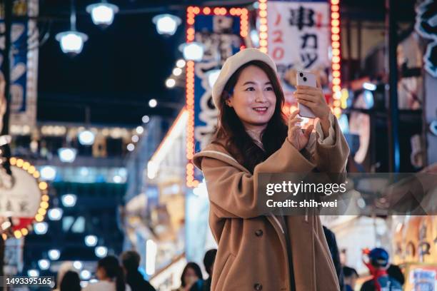 eine asiatische frau besichtigt die einkaufsstraße tsutenkaku in osaka, japan. abends nutzt sie ihr smartphone, um fotos von tsutenkaku und der umliegenden landschaft aufzunehmen, während die leuchtenden lichter im hintergrund leuchten. - osaka shinsekai food stock-fotos und bilder