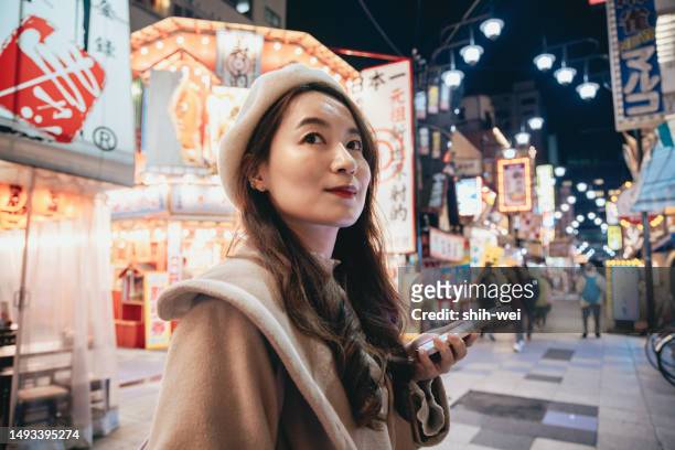 eine asiatische frau besichtigt die einkaufsstraße tsutenkaku in osaka, japan. nachts navigiert sie mit ihrem handy zu ihren gewünschten zielen, sucht nach lokaler küche und touristischen informationen. - osaka shinsekai food stock-fotos und bilder
