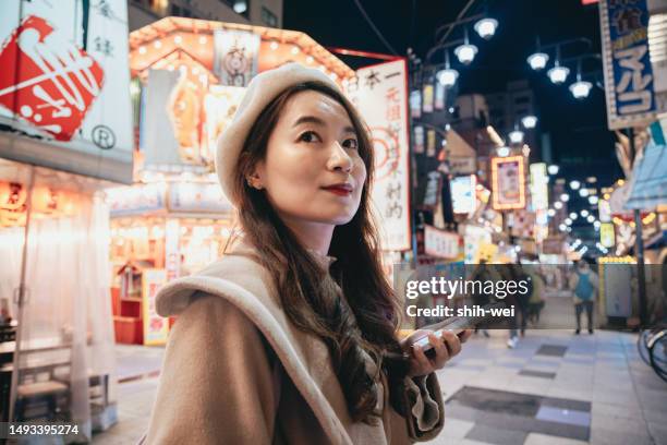 an asian woman is sightseeing at the shopping street of tsutenkaku in osaka, japan. during the night, she uses her mobile phone to navigate to her desired destinations, search for local cuisine and tourist information. - osaka shinsekai food stock pictures, royalty-free photos & images