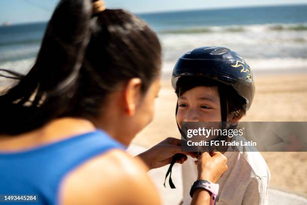 mother helping son to put sports helmet on a beach coastline - roupa desportiva de protecção imagens e fotografias de stock