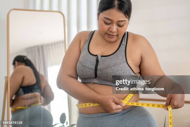 plus size female holds measures tape check her waist size. - woman-looking-into-mirror-full-body stock pictures, royalty-free photos & images