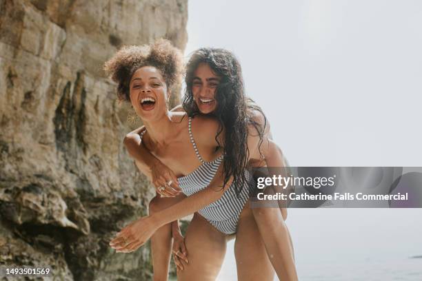 a woman gives a female friend a piggy back on the beach. - strandparty stock-fotos und bilder