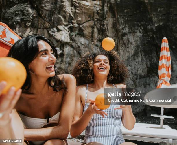 two beautiful woman juggle oranges on a sunny beach - friend mischief stock pictures, royalty-free photos & images