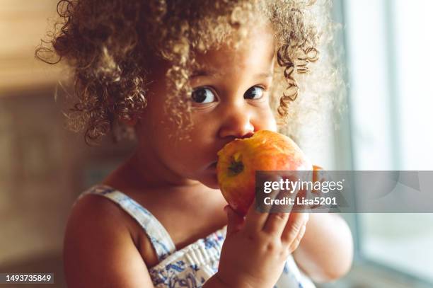 cute mixed race toddler girl eating a fresh red apple, healthy eating at home - kids eating stock pictures, royalty-free photos & images