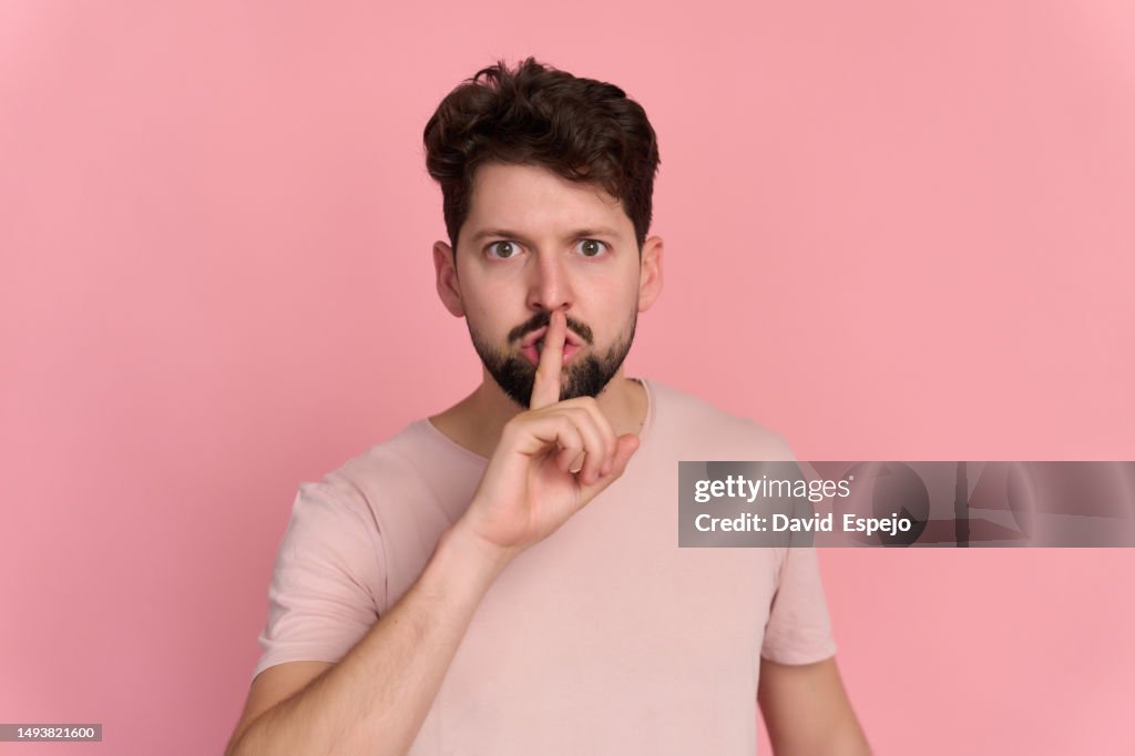 Man on pink background gesturing silence with finger over mouth