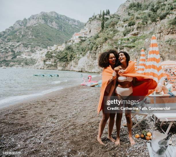two young woman stand on a beach, sharing a large orange beach towel. a view of positano, italy, is visible behind them. - strand liegen stock-fotos und bilder