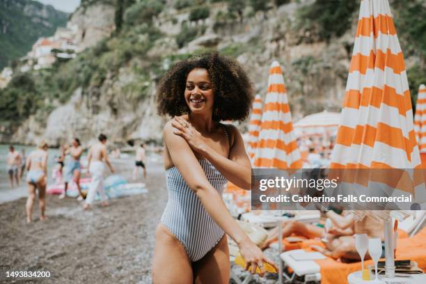 a beautiful young woman applies spray suncream on a sunny beach - badkleding stockfoto's en -beelden
