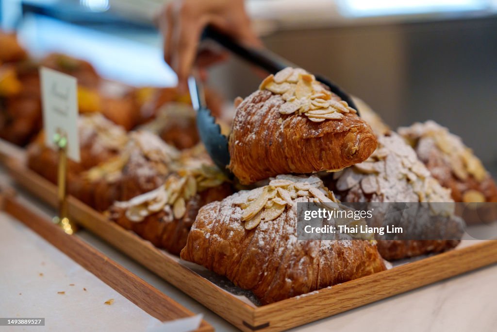 Cropped image of an Asian woman using tongs to pick up an almond croissant from a bakery store