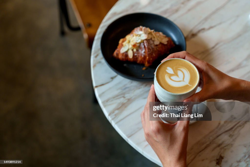 Over the shoulder view of an Asian woman enjoying her breakfast, a croissant with a cup of coffee in a cafe during the morning