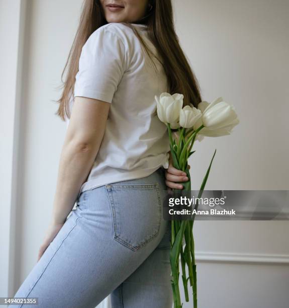 young happy smiling woman holding bunch of spring white tulip flowers. plus size woman wearing blue jeans and white t-shirt at home. self-love and self-confidence concept. - hair love 個照片及圖片檔