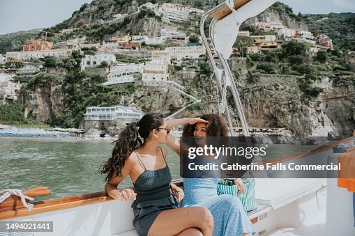 Two woman enjoy a journey on a boat, with Positano, Italy, in the background. One woman wears sunglasses, the other shields her eyes from the bright light with her hands. Two woman enjoy a journey on a boat, with Positano, Italy, in the background. One woman wears sunglasses, the other shields her eyes from the bright light with her hands.