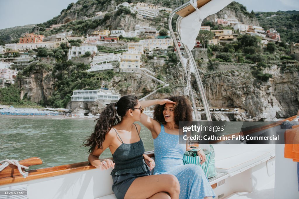Two woman enjoy a journey on a boat, with Positano, Italy, in the background. One woman wears sunglasses, the other shields her eyes from the bright light with her hands.