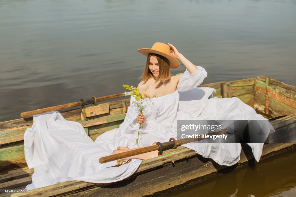 Blonde woman enjoying vacation on the lake. White linens in old fishing boat with oar. Bed sheets. Summer flowers