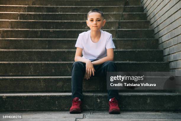 slim pre-adolescent boy in white t-shirt, black pants and red sneakers sitting on concrete stairs outside - schlank stock-fotos und bilder