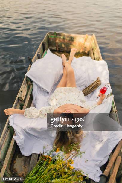 blonde woman in white dress enjoying vacation on the lake. white linens in old fishing boat with oar. bed sheets. summer relaxation - lazy river stock pictures, royalty-free photos & images