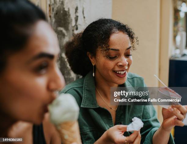 two woman sit on an outdoor stoop eat  ice-cream - one uses a tub and the other has wafer cone. - yoghurt tub stock pictures, royalty-free photos & images