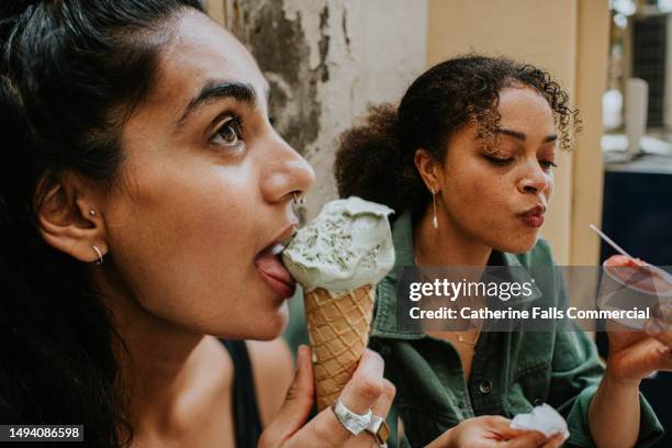two woman sit on an outdoor stoop eat  ice-cream - one uses a tub and the other has wafer cone. - yoghurt tub stock pictures, royalty-free photos & images