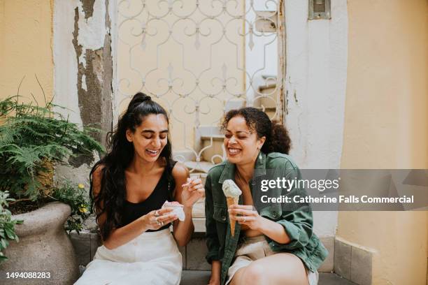 two woman sit on a stoop in an alley way and enjoy an ice cream together - gelato stock-fotos und bilder