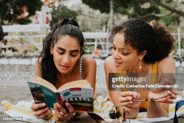 two woman lie on their stomachs on a beach. they read a guide / travel book together - jet lag stockfoto's en -beelden