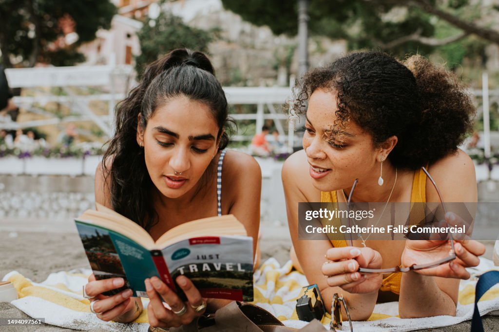 Two woman lie on their stomachs on a beach. They read a guide / travel book together