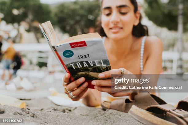 a woman lies on her stomach on a beach, and reads a travel guide - advice woman travel traveling stock pictures, royalty-free photos & images