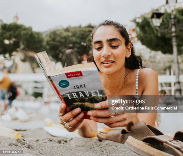 a woman lies on her stomach on a beach, and reads a travel guide - indian tour guide stock pictures, royalty-free photos & images