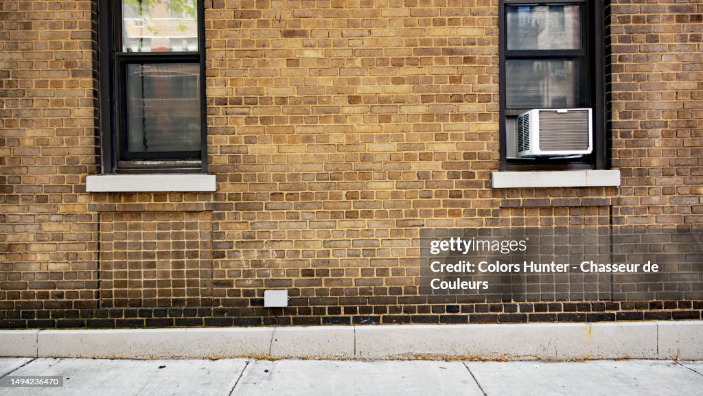 Patinated brown brick wall with windows, air conditioner and cement sidewalk in Manhattan, New York State, United States