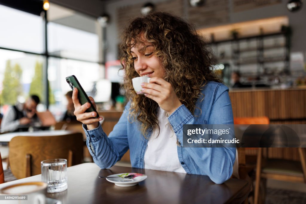 Jeune femme heureuse utilisant son téléphone portable et dégustant un café au café