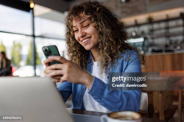 young smiling woman using mobile phone while working on a laptop at a cafe - mobile banking stock pictures, royalty-free photos & images