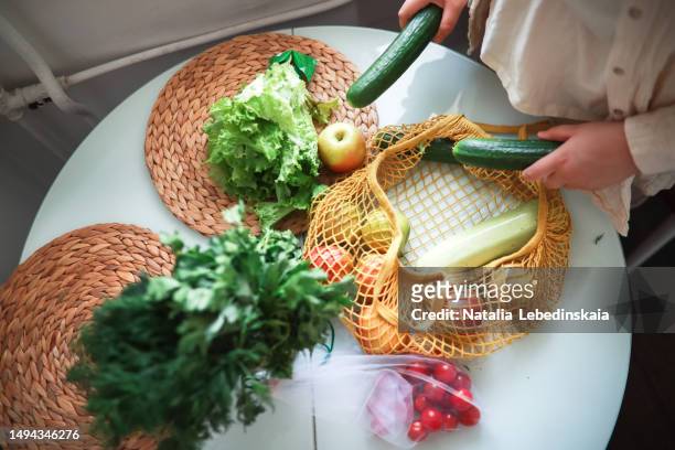 from bag to table: viewed from above, woman unveils range of fresh organic vegetables, zucchini, and cucumbers, plucked from mash bag on sunlit kitchen table, highlighting the principles of zero waste and responsible environmental practices. - groene salade stockfoto's en -beelden