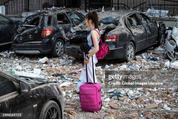 Girl with a suitcase stands next to cars damaged or wrecked by the rubble of a residential building hit by a downed kamikaze drone on May 30, 2023 in...
