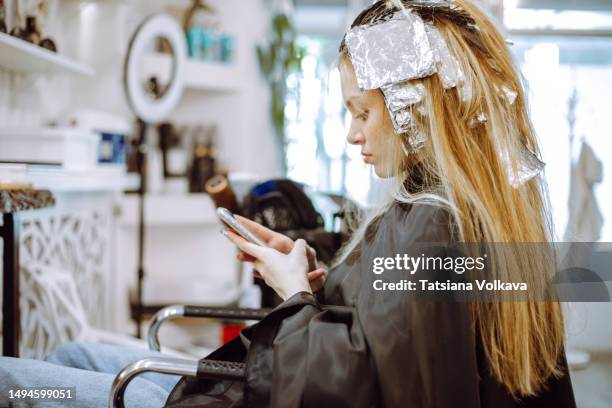 side view of teenage girl sitting and using mobile phone during hair coloring procedure at hairdresser shop - teenagers cheveux colorés photos et images de collection